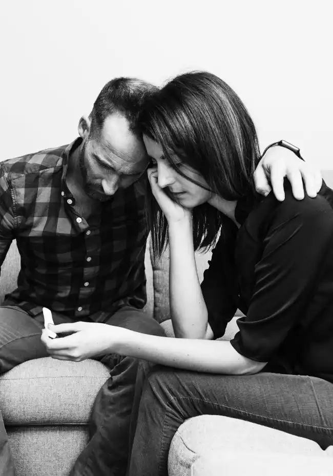 Ein Mann und eine Frau sitzen an einem Tisch und halten sich an den Händen. Die Frau wirkt traurig.